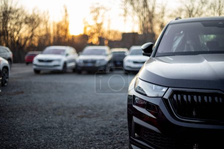 Photo for Partial front view of new black modern family SUV. Visible front LED light and part of gloss black grille and bumper. Out-of-focus cars and trees in the background. Sunset light. Parking on gravel. - Royalty Free Image