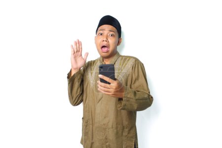 Photo for Asian muslim man holding mobile phone raising hand show shocked expression  isolated on white background, looking at camera - Royalty Free Image
