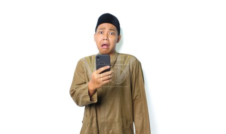 Photo for Shocked asian muslim man holding mobile phone looking at camera with scared expression isolated on white background - Royalty Free Image