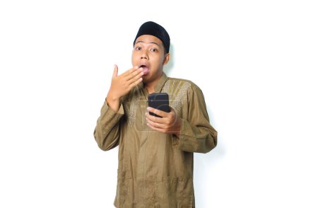 Photo for Asian muslim man holding mobile phone covering his mouth show shocked expression  isolated on white background, looking at camera - Royalty Free Image