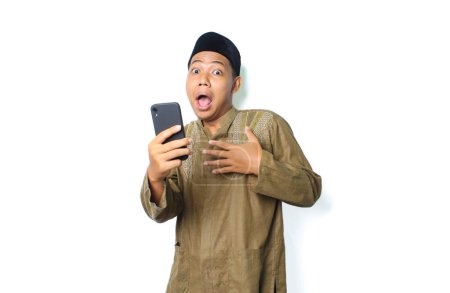 Photo for Asian muslim man holding mobile phone touching his chest show shocked expression  isolated on white background, looking at camera - Royalty Free Image