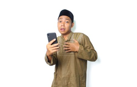 Photo for Asian muslim man holding mobile phone touching his chest show shocked expression  isolated on white background, looking at camera - Royalty Free Image
