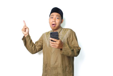 Photo for Shocked asian muslim man holding mobile phone pointing finger above isolated on white background, looking at camera - Royalty Free Image