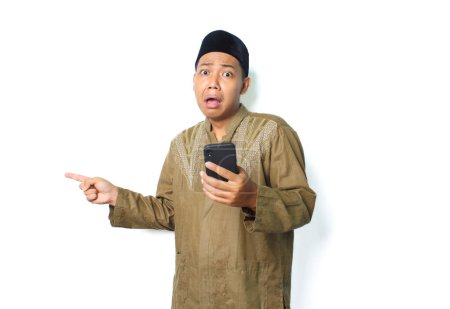 Photo for Shocked asian muslim man holding mobile phone pointing finger beside isolated on white background, looking at camera - Royalty Free Image