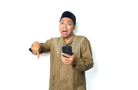 Photo for Shocked asian muslim man holding mobile phone pointing finger down isolated on white background, looking at camera - Royalty Free Image