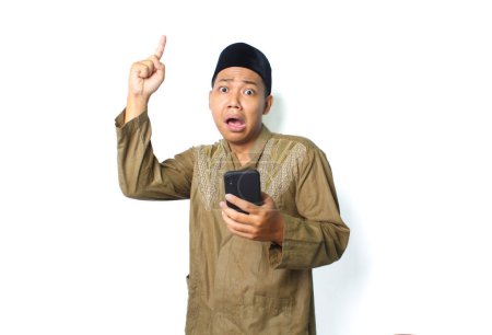 Photo for Shocked asian muslim man holding mobile phone pointing finger above isolated on white background, looking at camera - Royalty Free Image