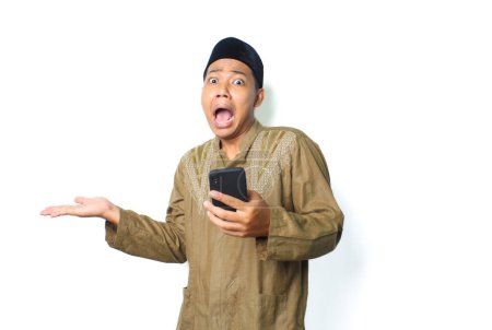 Photo for Shocked asian muslim man presenting copy space with holding mobile phone, looking at camera, isolated on white background - Royalty Free Image