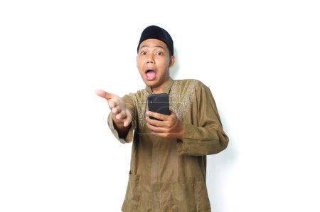 Photo for Shocked asian muslim man wearing islamic dress offering handshake with holding smartphone isolated on white background, looking at camera - Royalty Free Image
