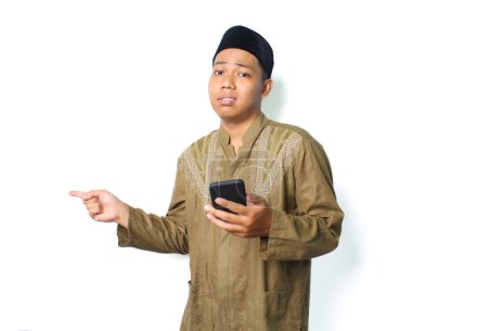 Photo for Anxious asian muslim man holding mobile phone with pointing to beside show worry expression isolated on white background - Royalty Free Image