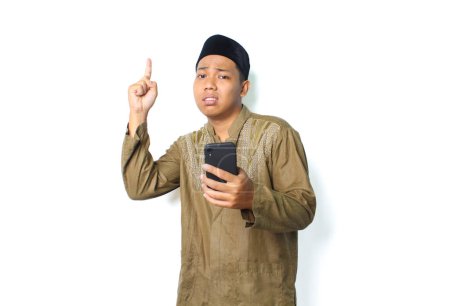 Photo for Anxious asian muslim man holding mobile phone with pointing to above show worry expression isolated on white background - Royalty Free Image