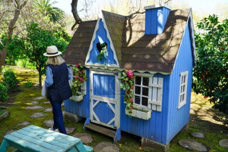 Téléchargez les photos : Une grand-mère impatiente attend de jouer avec ses petits-enfants dans le charmant jardin à côté de la maison de poupée en bois bleue - en image libre de droit
