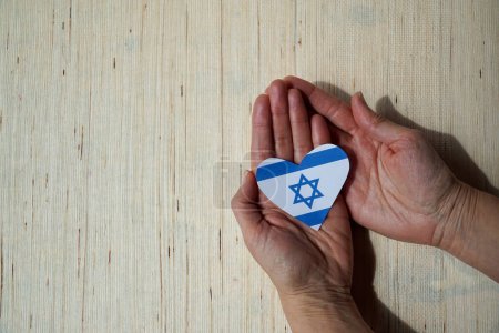 Téléchargez les photos : Deux mains bercent tendrement un drapeau israélien en forme de cœur, sur fond de bois clair. - en image libre de droit