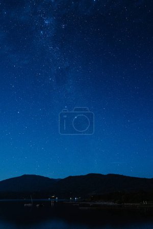 Foto de Noche estrellada iluminando el cielo azul con una vista de la Vía Láctea sobre un pueblo junto al lago. - Imagen libre de derechos