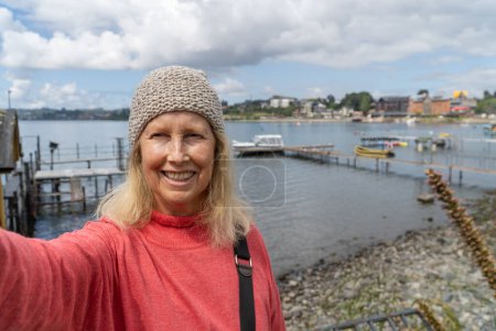 Senior tourist woman capturing a selfie in a picturesque town beside a serene lake. Site seeing in puerto varas, Chile.