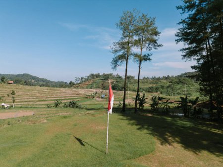 Indonesian flag waving proudly against a backdrop of fields, rice paddies, and lush trees, symbolizing national pride and natural beauty.