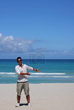 Young man with tan and sunglasses presenting something in the copy space area of the beach landscape.