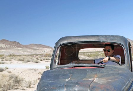 Young man wearing sunglasses sitting in an abandoned car wreck in Death Valley