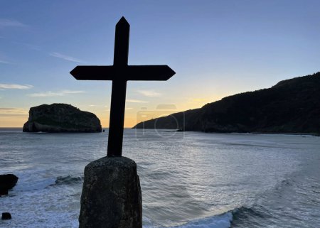 Oceanfront at San Juan de Gaztelugatxe in Spain during sunrise with a cross in the foreground