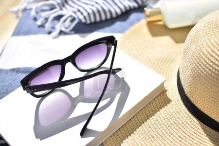 Téléchargez les photos : Lunettes de soleil modernes, un livre, crème solaire, un chapeau de paille se trouvent sur une serviette de plage. Vacances d'été à la plage. - en image libre de droit