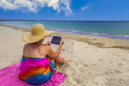Foto de Female with pareo holding tablet by an empty sandy coast with calm waves. Day view of woman with naked legs wearing a straw hat & colorful kaftan beachwear, sitting on a beach towel by the sea. - Imagen libre de derechos