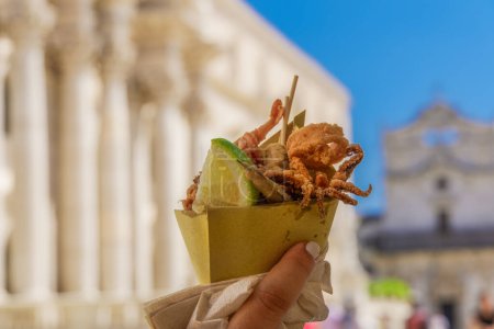 Photo for Holding Italian street fish food on a cone. Day view of a female hand that holds a greasy paper cone with seafood including pan fried squid, small fish & lime against blurred background in Ortygia. - Royalty Free Image