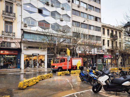 Foto de Tesalónica, Grecia - 3 de abril de 2023: Bomberos con abrigos protectores amarillos cortando ramas de árboles en el centro de la ciudad durante un clima lluvioso muy ventoso. - Imagen libre de derechos