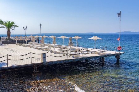 Téléchargez les photos : Terrasse de bronzage en bois vide avec chaises longues et parasols au bord d'une mer bleue calme sous le soleil éclatant en été. - en image libre de droit