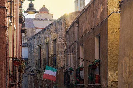 Callejón de la ciudadela de Cagliari con edificios tradicionales con balcones de hierro y bandera italiana ondeando en la isla de Cerdeña, Italia.