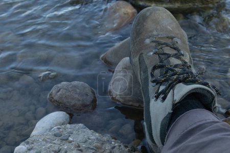 Primer plano de una bota de senderismo que descansa sobre rocas en un río claro, mostrando aventura al aire libre y naturaleza.