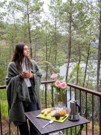 Téléchargez les photos : Portrait image d'une belle femme caucasienne tenant et buvant du café chaud, debout sur le balcon et regardant le lac et la nature verte. - en image libre de droit