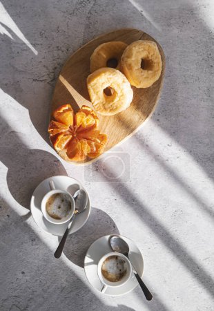 Foto de Trío de rosquillas sobre tabla de madera con mandarina y dos tazas de café, bañadas en luz natural. - Imagen libre de derechos