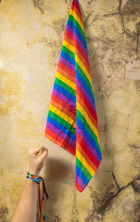 Mano de una mujer con puño cerrado y brazalete de arco iris, fondo bandera lgbti, luchando por la tolerancia y el amor.