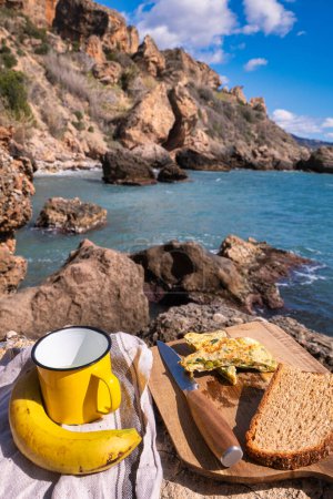 Omelet on wooden board with bread and a cup. In the background rocks on the seashore