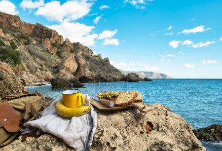 Food of a hiker. Omelet on wooden board bread and a cup, next to it a backpack. In the background cliffs in the sea,