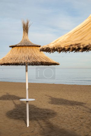 Parapluie en paille au premier plan sur une plage au coucher du soleil, environnement calme et serein sans personnes.