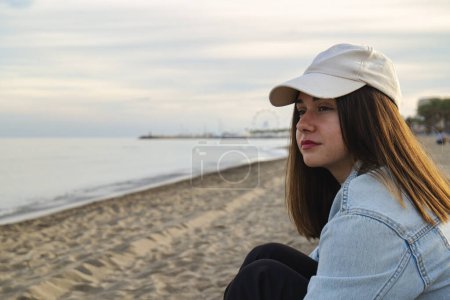 Chica caucásica con pelo largo y gorra mirando serenamente al horizonte en la orilla del mar, escena tranquila de paz y serenidad.