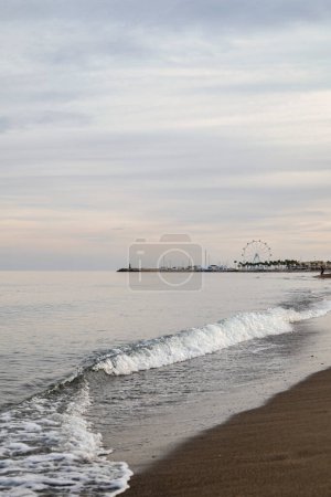Sea scenery, waves breaking on the fork of a beach, in the background a small harbor with a ferris wheel