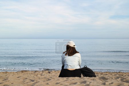 Mädchen mit Mütze sitzt am Ufer eines Strandes ohne Menschen, Gefühl von Gelassenheit und Einsamkeit.
