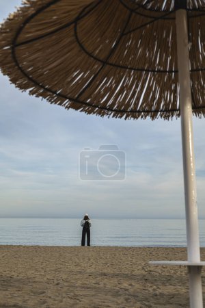 Sombrilla de playa en primer plano fuera de foco, en el fondo una chica en su espalda en la orilla del mar mirando al horizonte.
