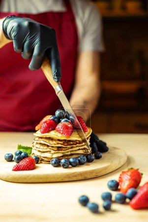 Mann schneidet Sahne-Pfannkuchen auf einem Holzbrett mit Erdbeeren und Blaubeeren, warmes Licht, Bar
