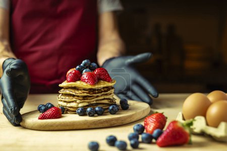 Mann in Schürze und schwarzen Handschuhen präsentiert Pfannkuchen mit Sahne, Erdbeeren und Blaubeeren auf einem Holzbrett, warmes Licht