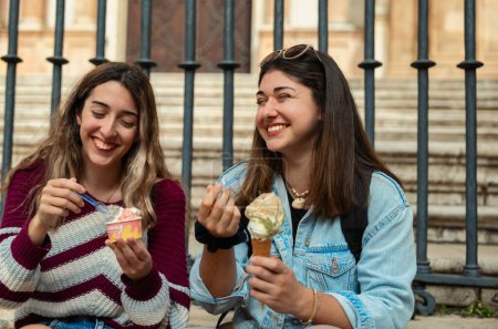 Two Caucasian girls sitting on a staircase eating ice cream and chatting with smiling faces