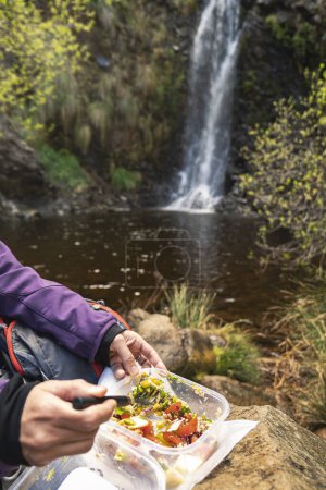 Unrecognizable woman eating from a tupper in the middle of nature, in the background a waterfall forming a pool of water, adventure and tourism.