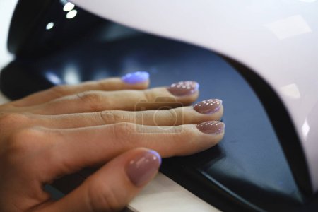 Téléchargez les photos : Femme séchant des ongles dans la lampe. Client avec vernis beige au salon de manucure attendant soigneusement lorsque ses ongles deviennent secs à la lumière UV. Poussée rapprochée - en image libre de droit