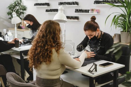 Photo for Doing manicure in salon. Female manicurist working on client nails in professional salon. Occupation concept. Customers at beauty green modern studio - Royalty Free Image