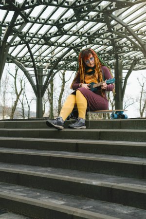 Foto de Mujer hipster libre divertido con rastas sentado en longboard en la carretera tocando ukelele guitarra a sus 30 años. hembra con pelo colorido brillante disfrutar de estilo de vida activo al aire libre, reproducir música - Imagen libre de derechos