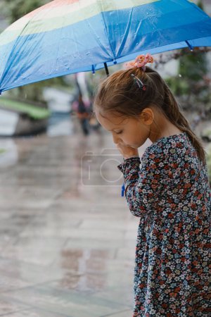 Téléchargez les photos : Enfant avec parapluie marchant sous la pluie dans la ville, fille heureuse avec parapluie coloré arc-en-ciel près de la maison, temps chaud d'été humide avec des flaques - en image libre de droit