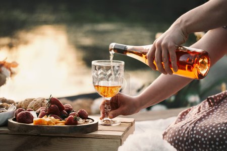 Photo for Rose wine on sunset picnic outdoors in summer. Glass of drink, fresh strawberries, cheese and pie for romantic eco style dinner. Woman pouring drink from the bottle - Royalty Free Image