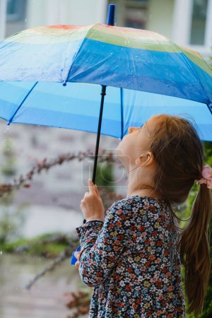 Foto de Niño con paraguas caminando bajo la lluvia en la ciudad, chica feliz con paraguas colorido arco iris cerca de la casa, clima cálido y húmedo de verano con charcos - Imagen libre de derechos