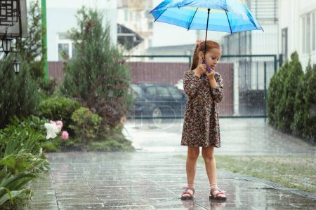 Téléchargez les photos : Enfant avec parapluie marchant sous la pluie dans la ville, fille heureuse avec parapluie coloré arc-en-ciel près de la maison, temps chaud d'été humide avec des flaques - en image libre de droit